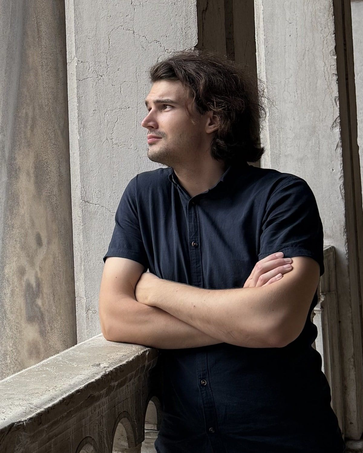 Ivan leaning on the stone balustrade of a loggia in Venice, arms crossed, looking off to one side.